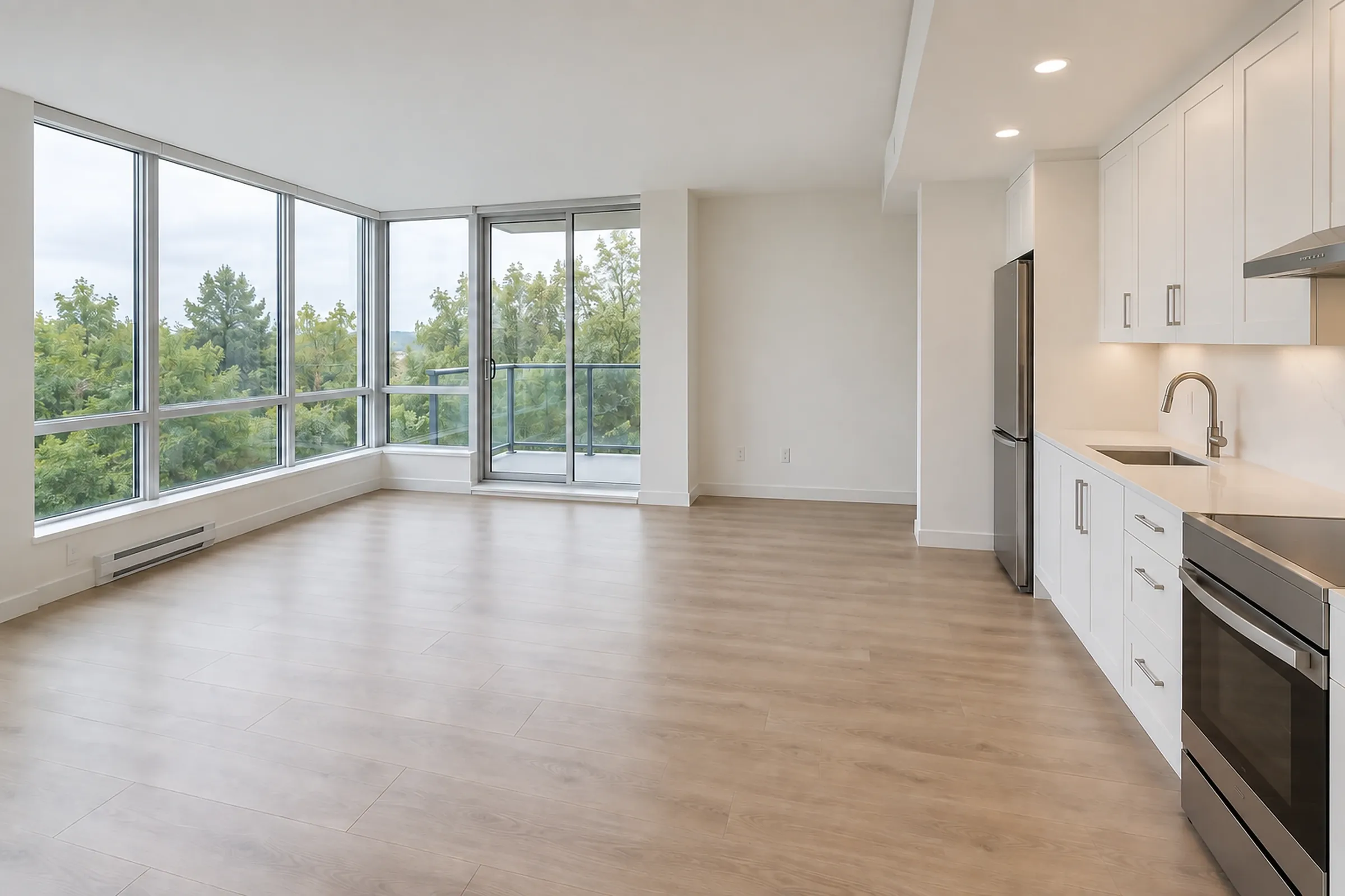 An empty, bright condo living area with corner windows, a balcony door and a galley kitchen